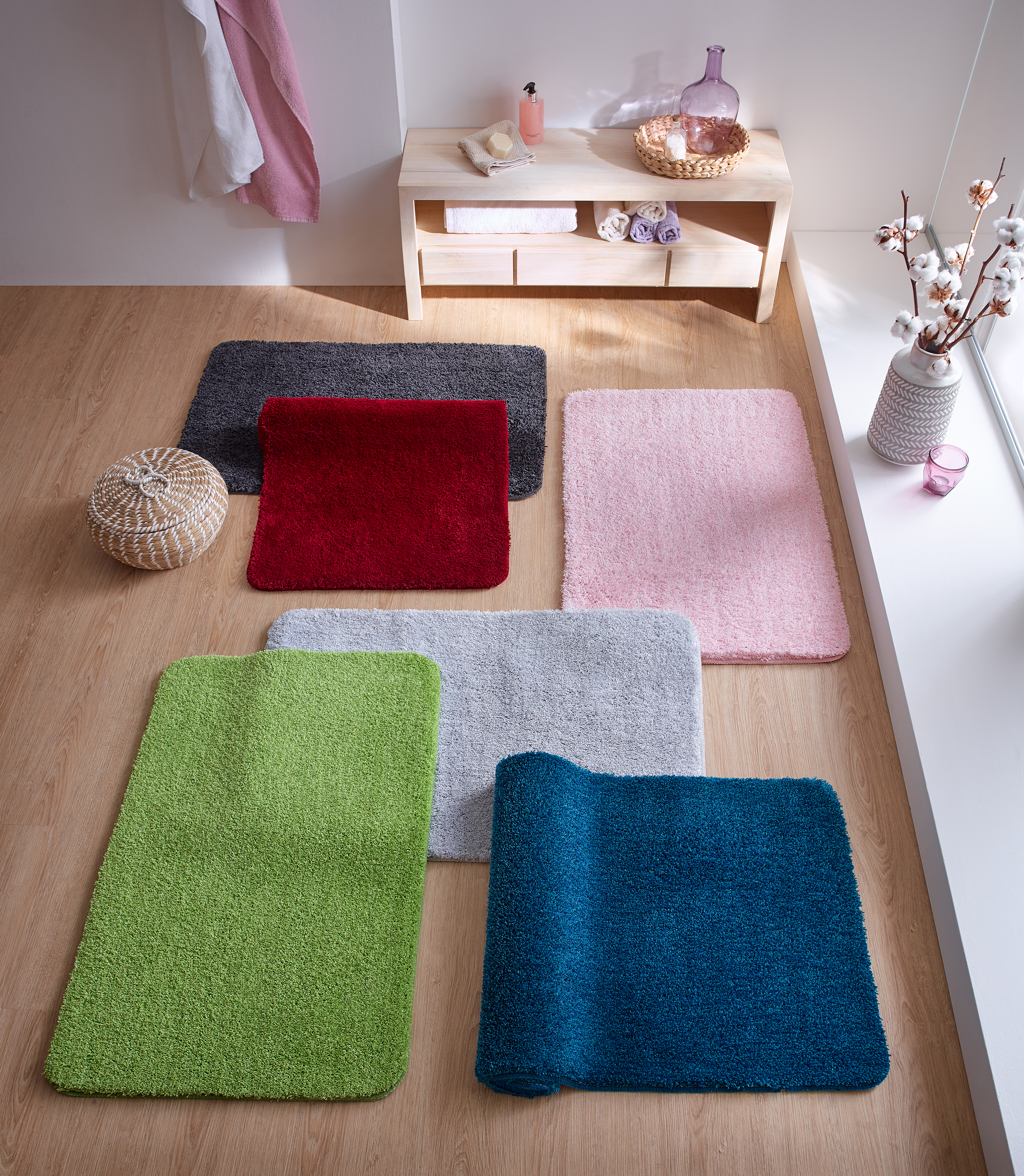 Bunte Badematten in verschiedenen Farben auf einem Holzboden, dekoriert mit einem Korb, Handtüchern und Accessoires.