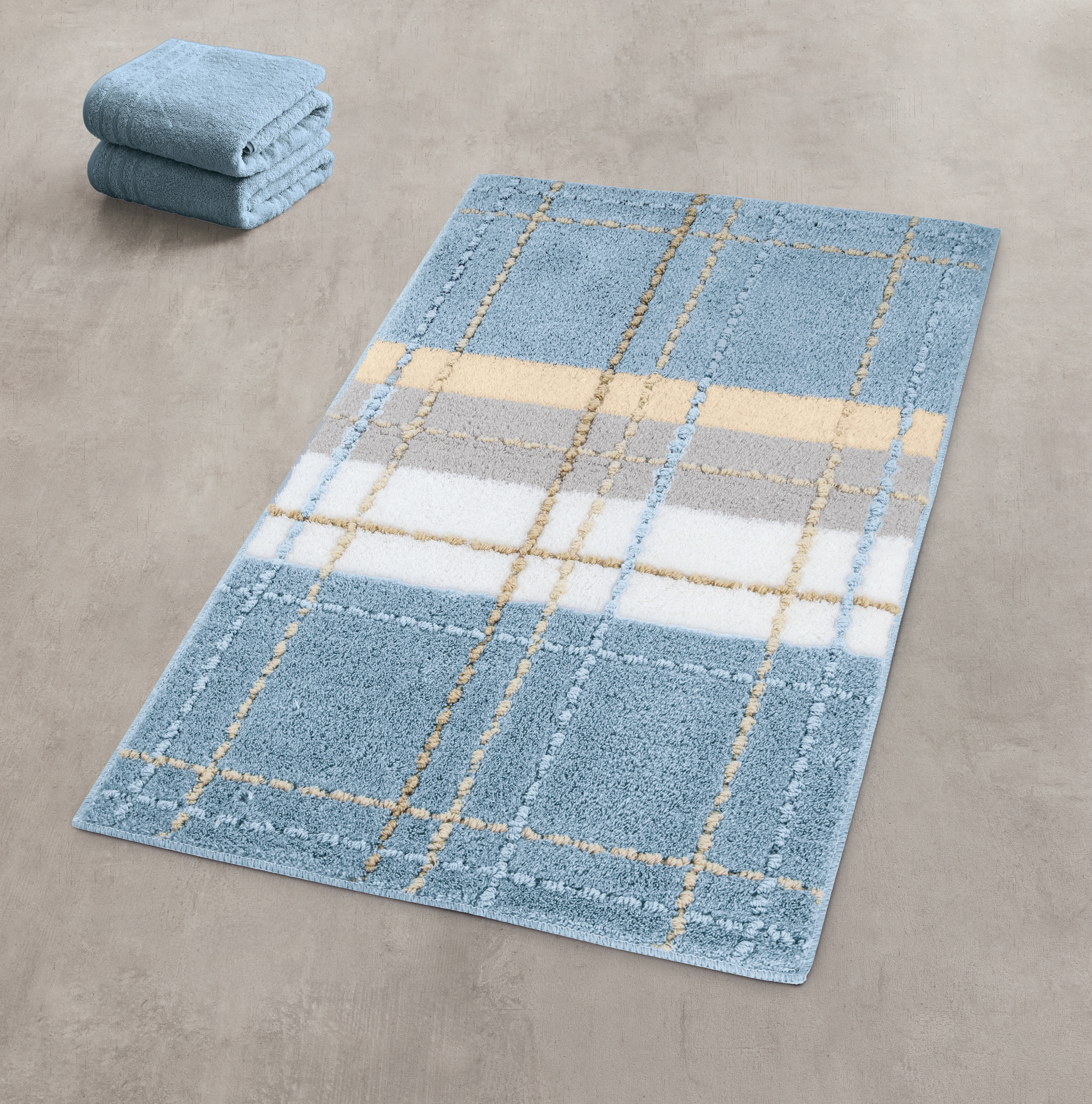 Weicher, blauer Badteppich mit Karomuster in Beige, Weiß und Grau, daneben gefaltete Handtücher auf grauem Boden.