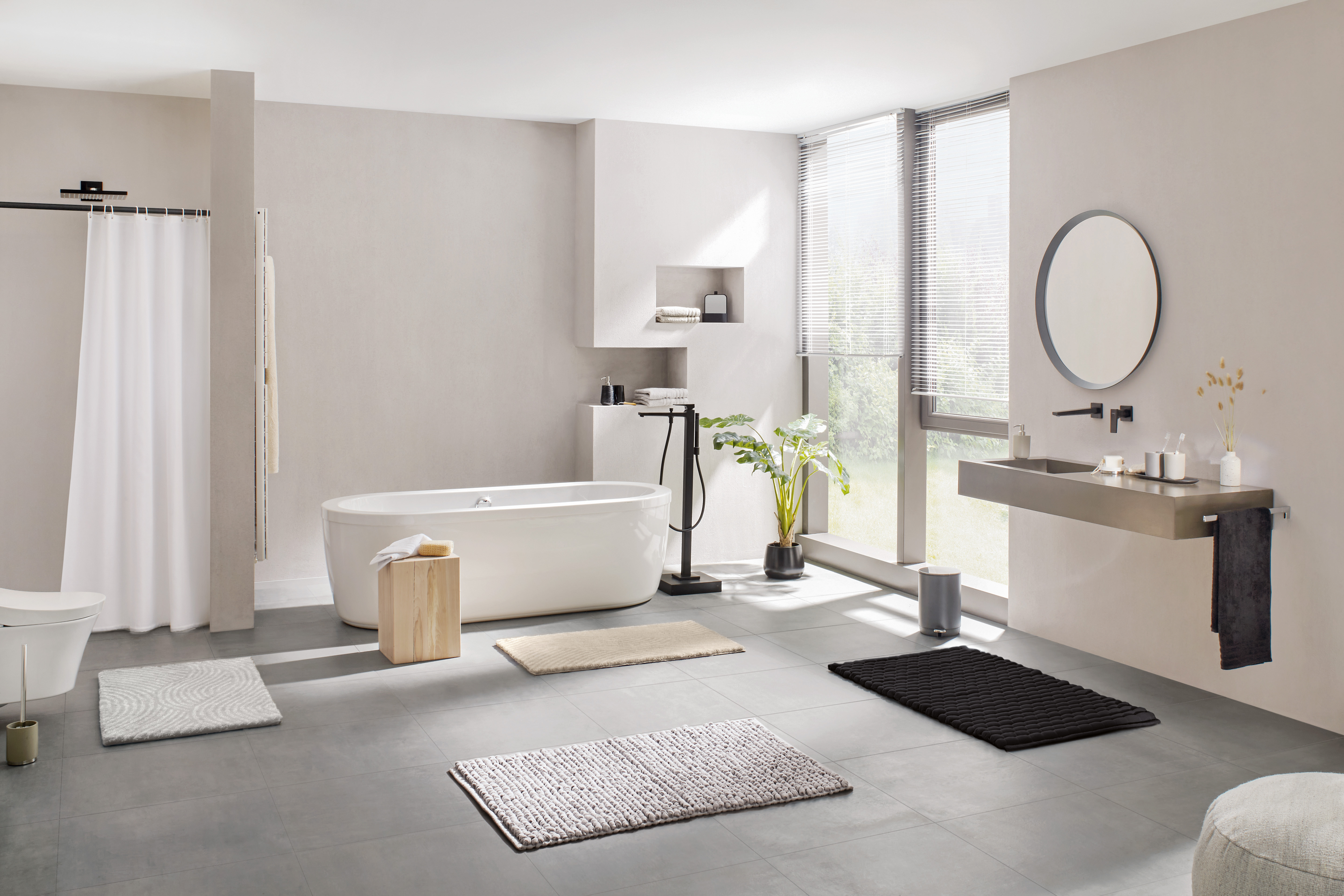 Modernes Badezimmer mit freistehender Badewanne, Waschbecken, Spiegel und verschiedenen Badteppichen in neutralen Farben.