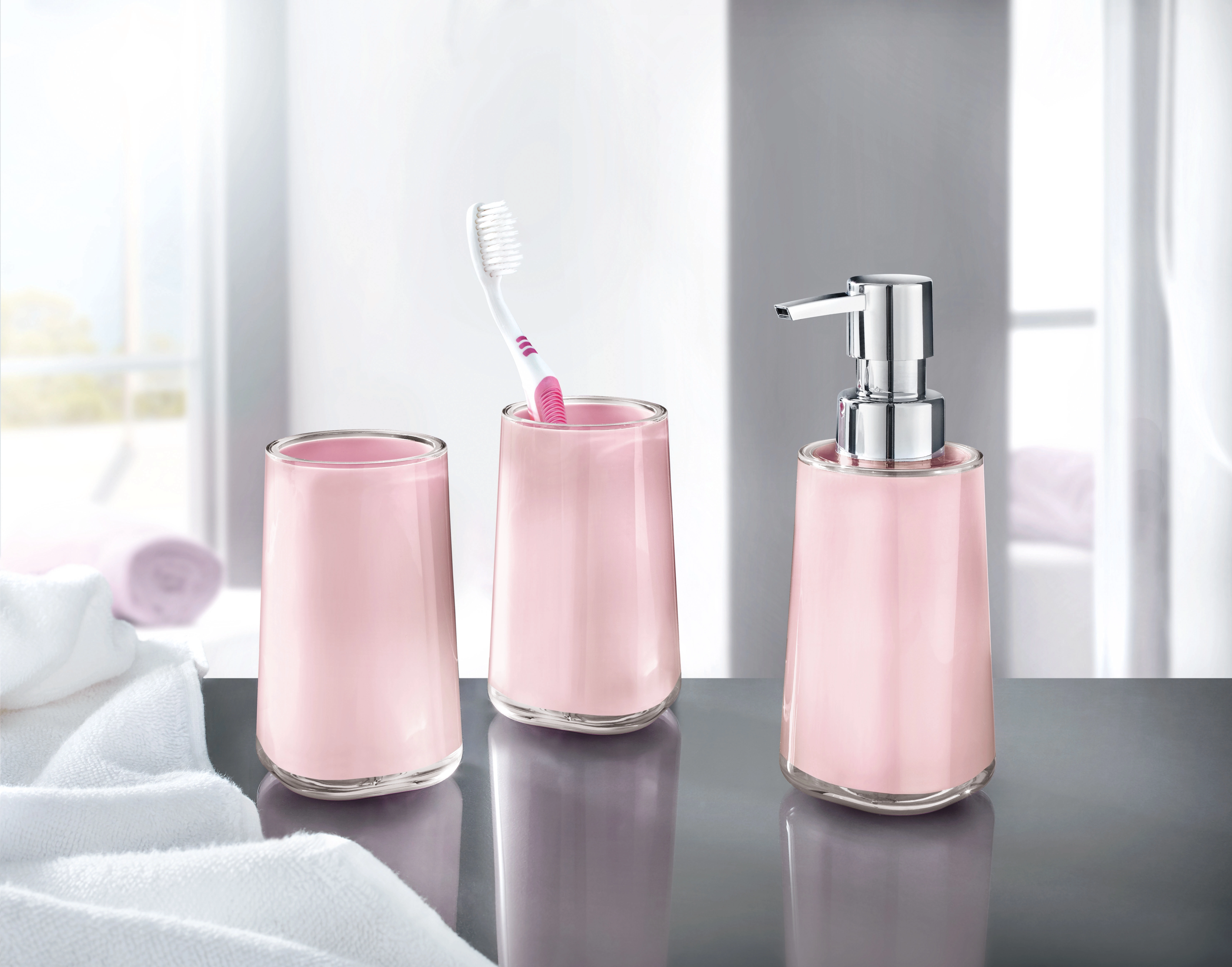 Rosa Badezimmer-Set mit Seifenspender, Zahnbürstenhalter und Becher auf grauer Oberfläche vor hellem Hintergrund.