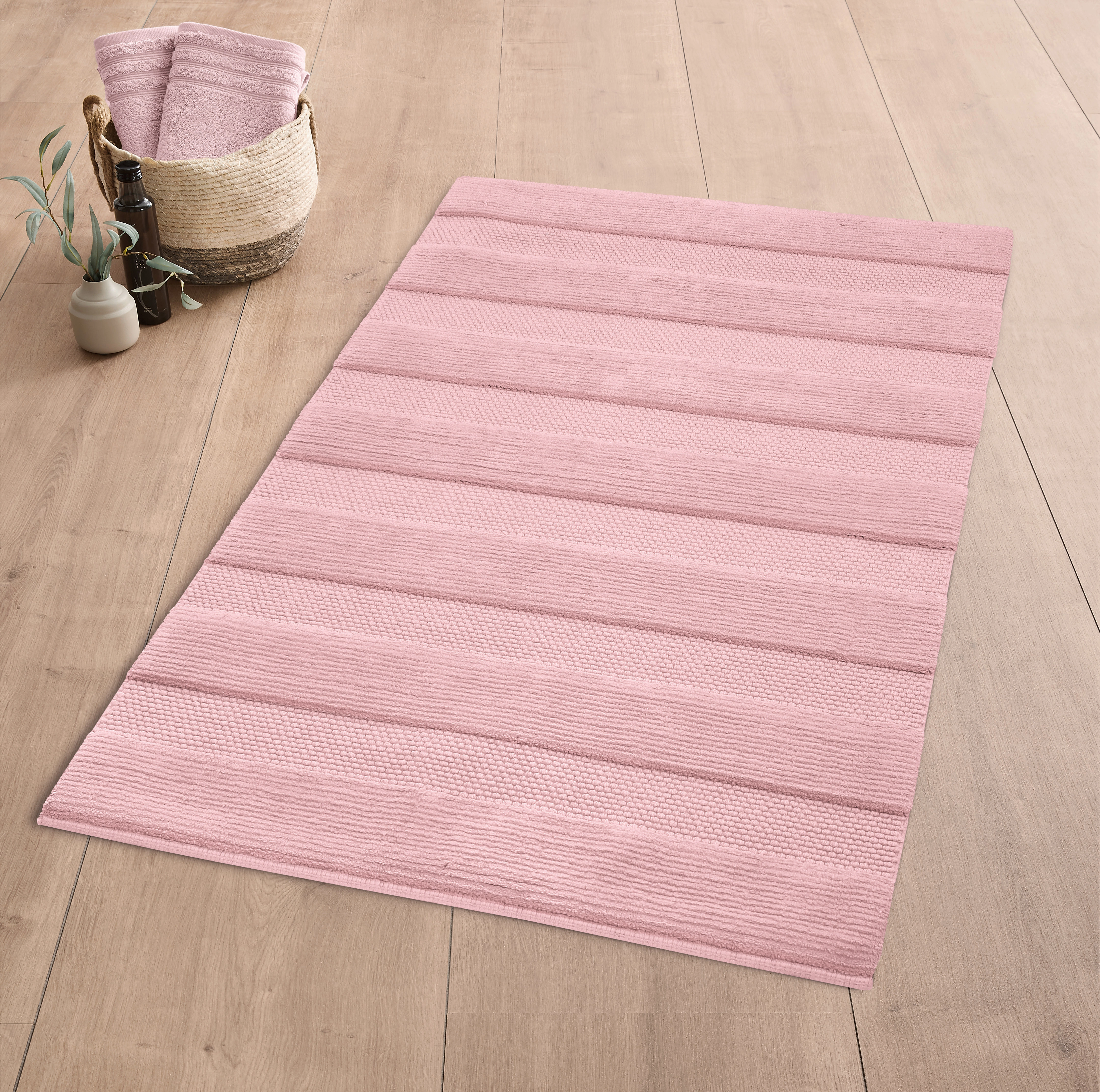 Rosa Badematte mit Streifenmuster auf Holzfußboden, dekoriert mit einem Korb und einer Pflanze.