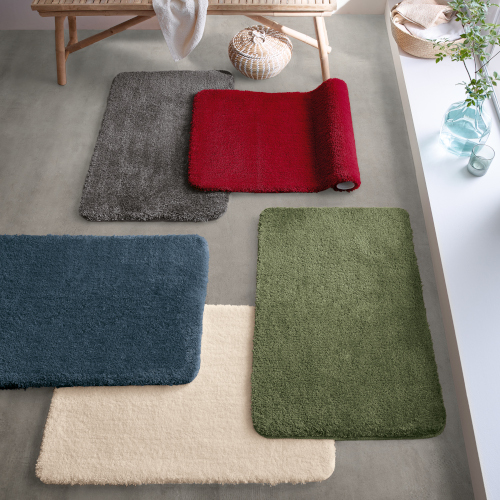 Bunte Badematten in verschiedenen Farben auf grauem Boden, dekoriert mit Pflanzen und einem Holzstuhl im Hintergrund.