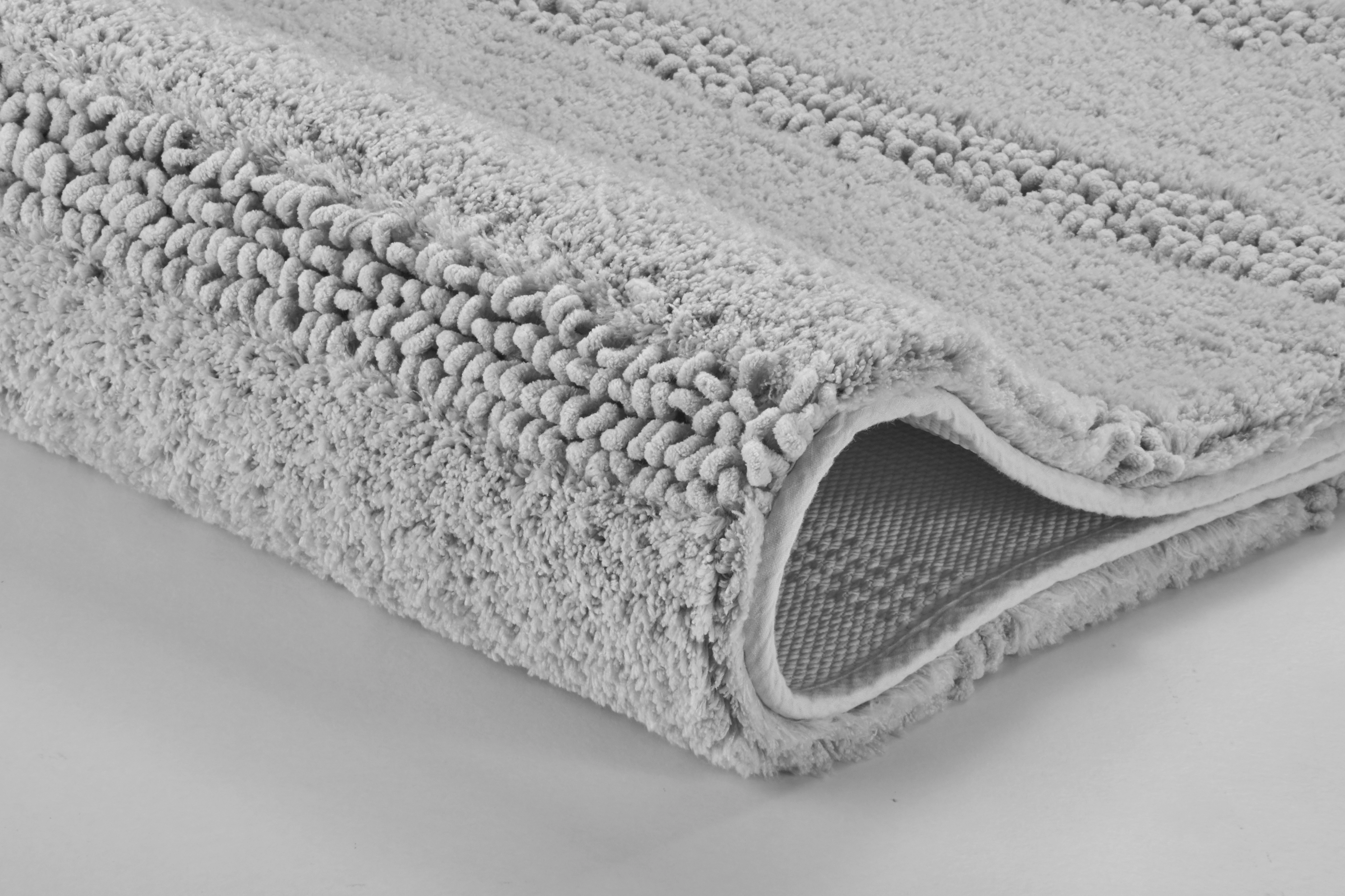 Grauer, flauschiger Badteppich mit rutschfester Unterseite, Nahaufnahme der Textur und Kanten.