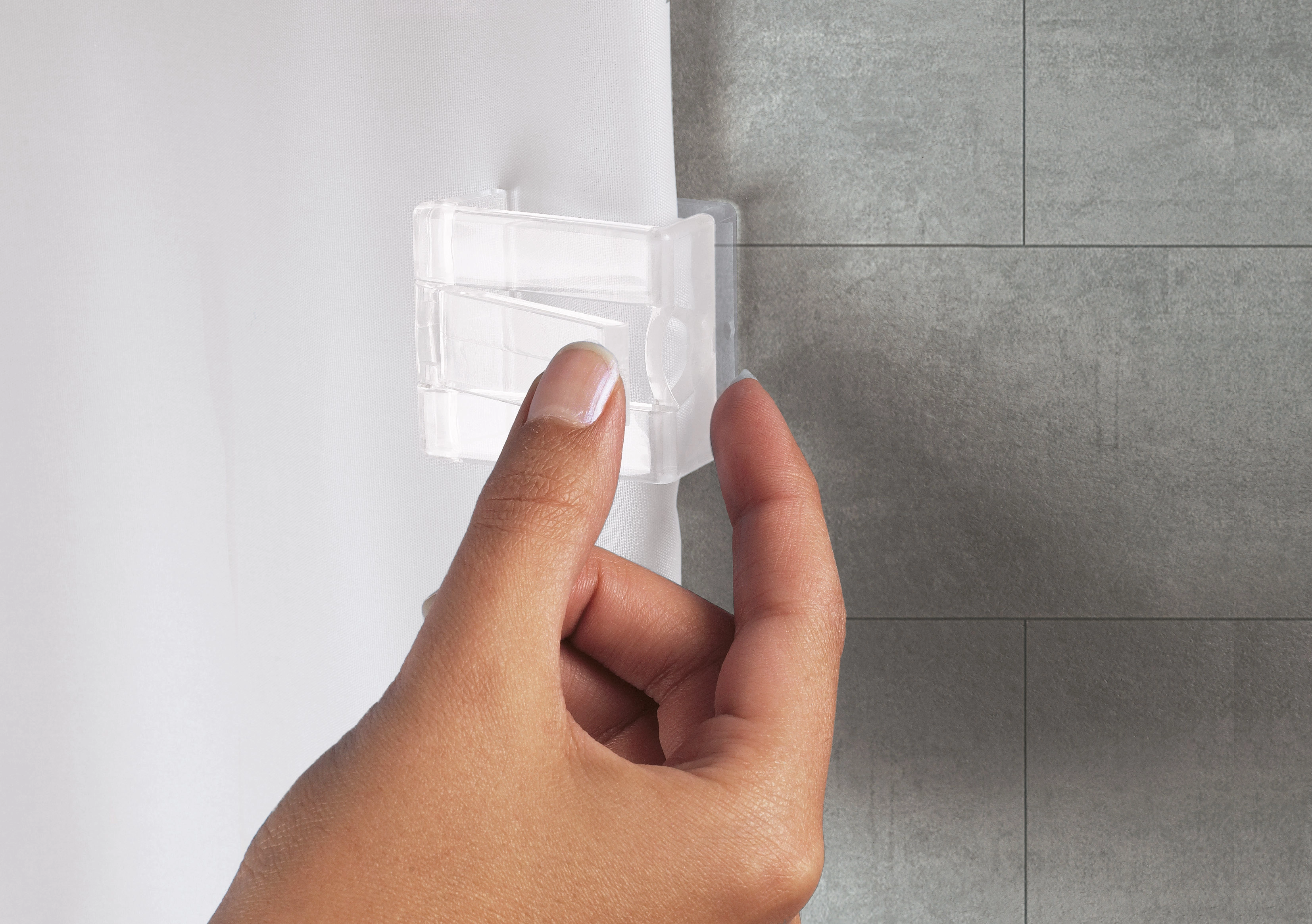 Hand befestigt transparente Duschvorhangklammer an weißem Vorhang vor grauer Fliesenwand.