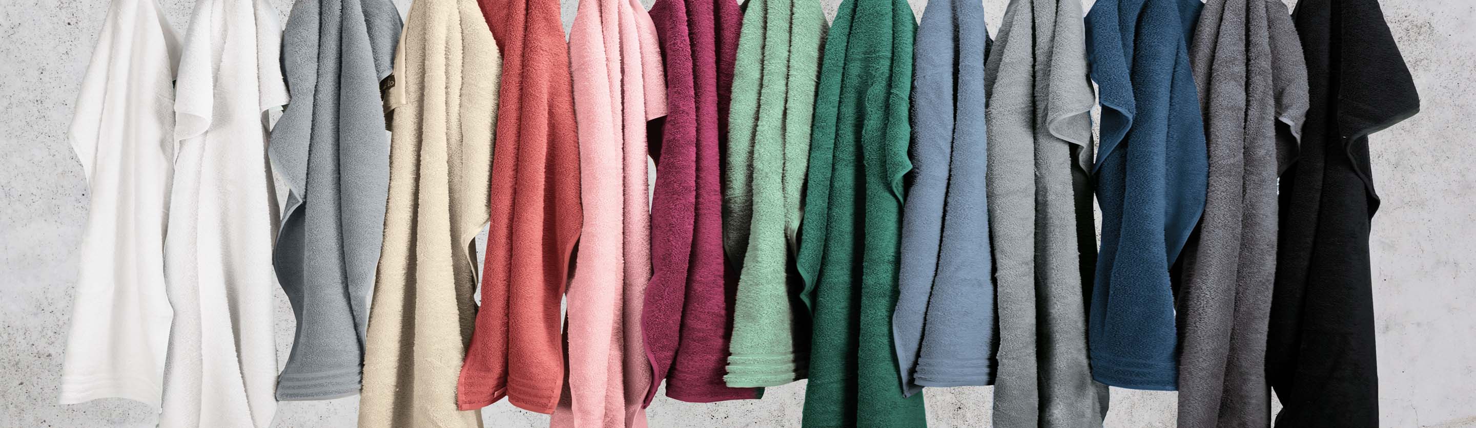 Bunte Handtücher in verschiedenen Farben, ordentlich nebeneinander aufgehängt, vor einer grauen Wand.