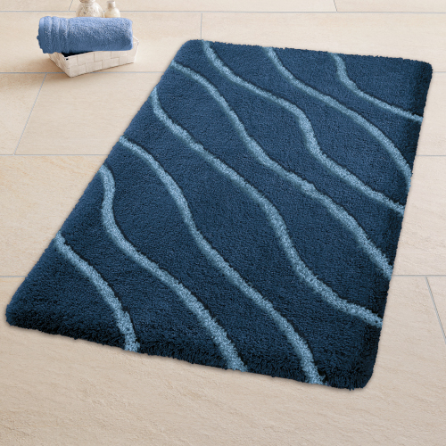 Dunkelblauer Badteppich mit wellenförmigem Muster auf beigem Fliesenboden, daneben ein Korb mit Handtüchern.