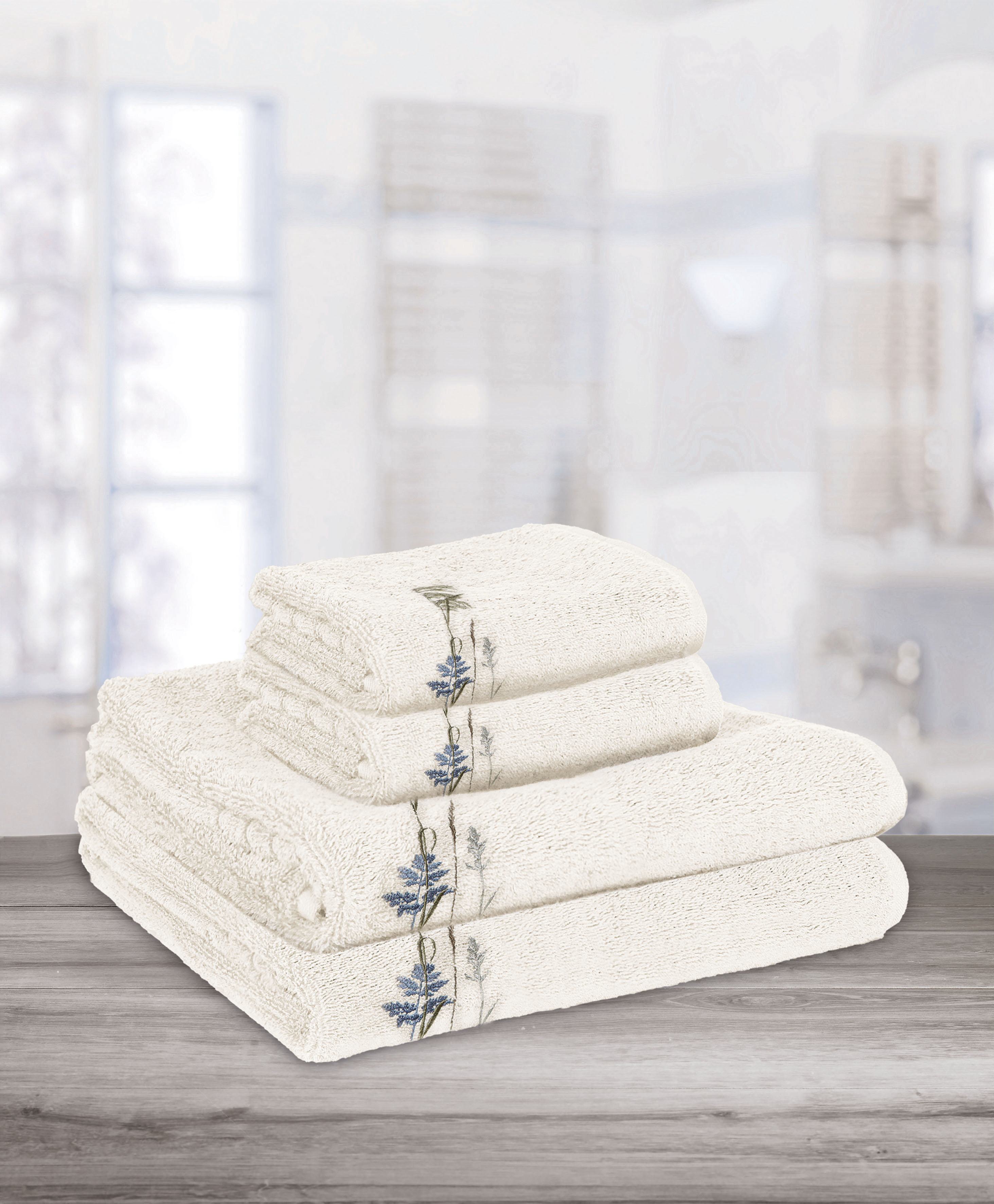 Weißes Handtuch-Set mit blauer Blumenstickerei, präsentiert auf einem Holztisch in einem hellen Badezimmer.
