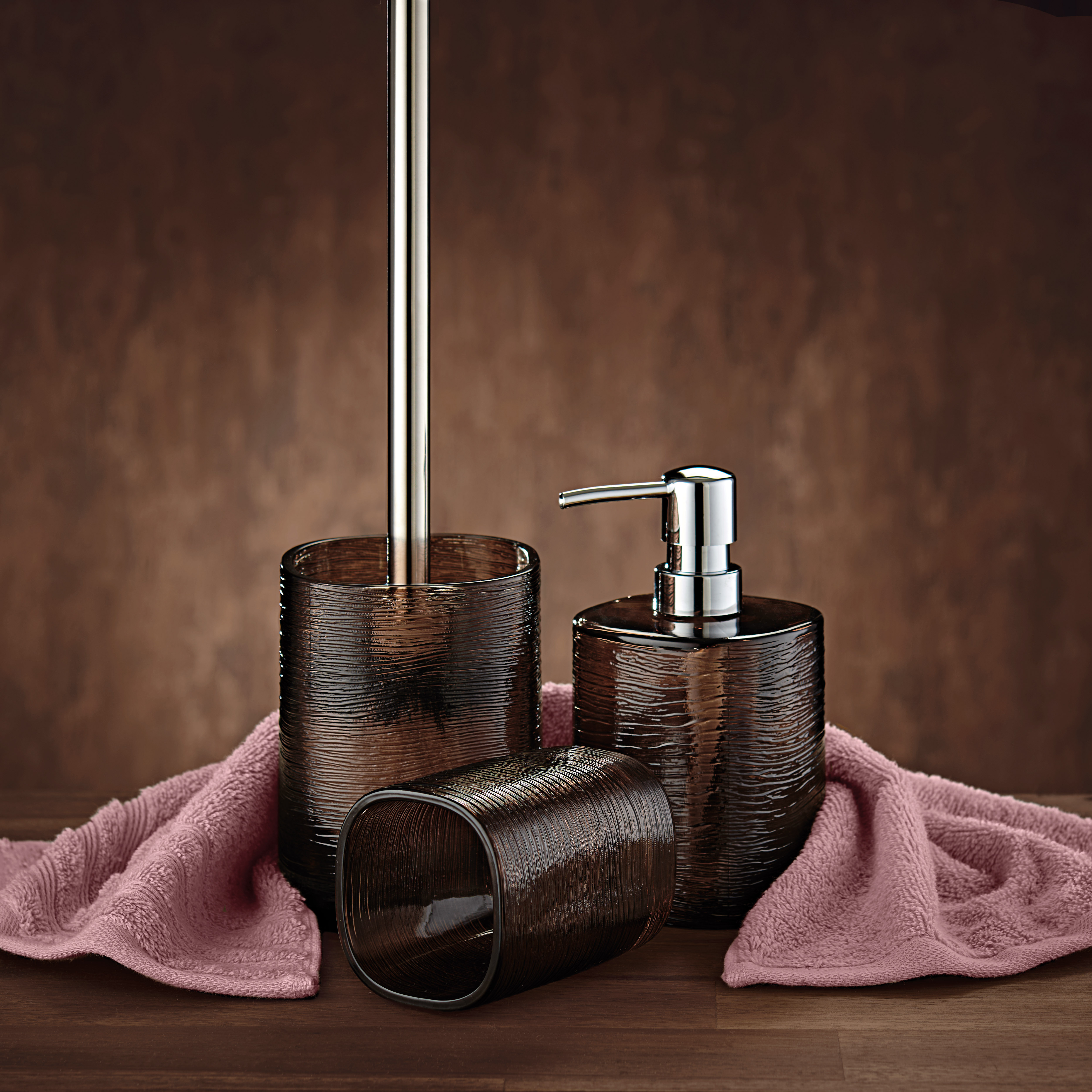 Braunes Bad-Accessoire-Set aus Seifenspender, Zahnbürstenhalter und WC-Bürste auf rosa Handtuch vor braunem Hintergrund.