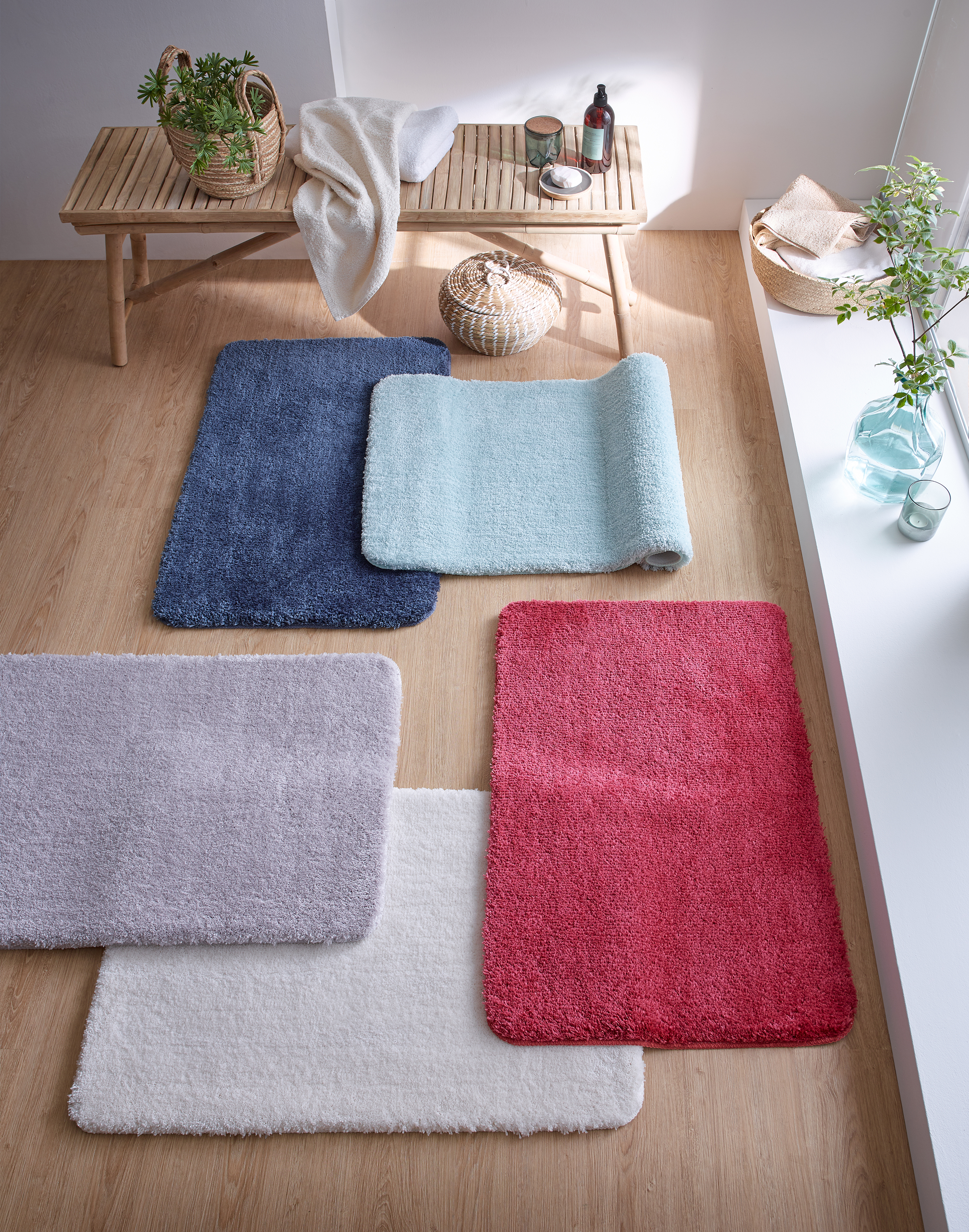 Bunte, flauschige Badematten in verschiedenen Farben auf einem Holzboden, dekoriert mit Pflanzen und Accessoires.