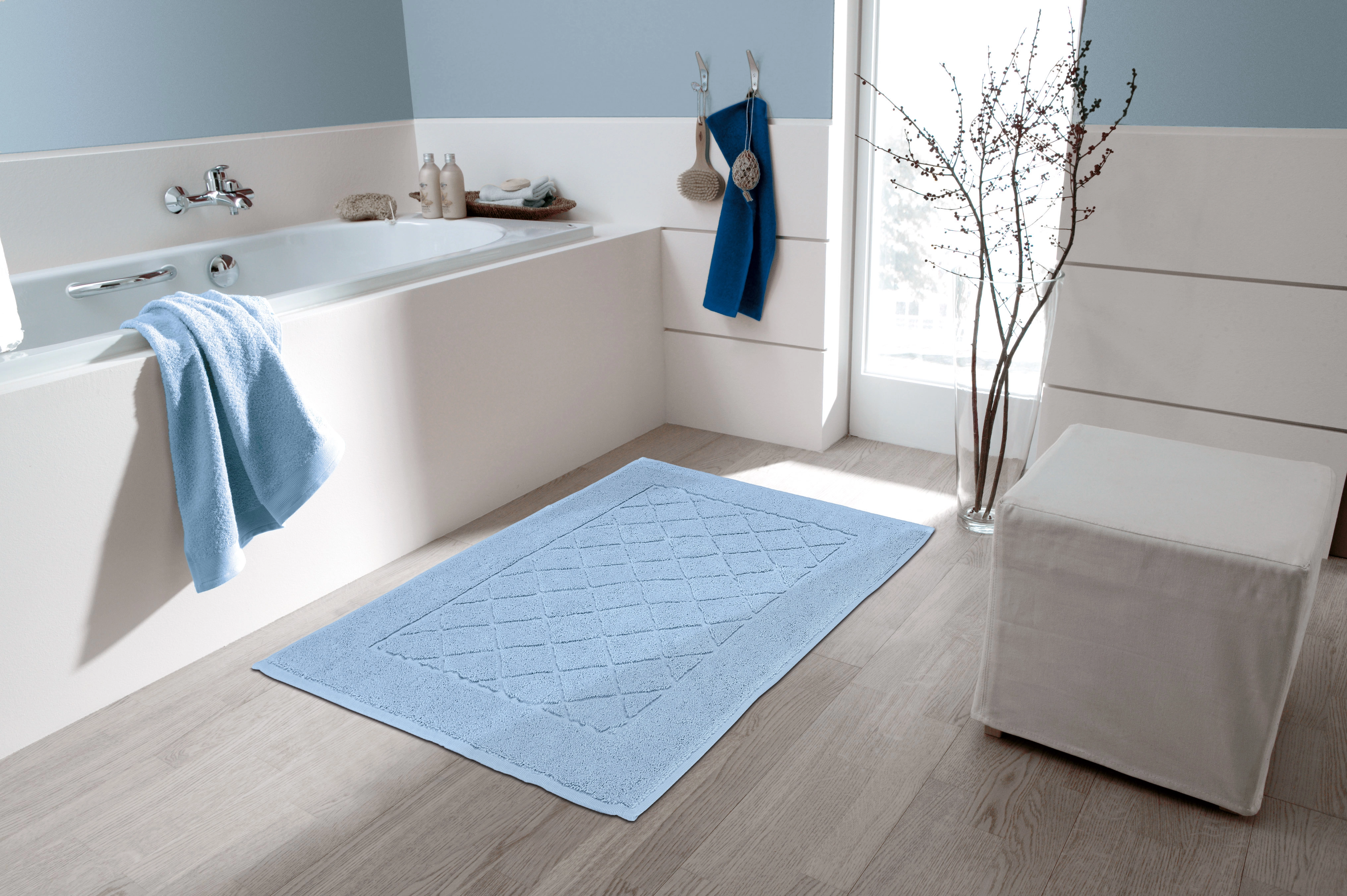 Helles, modernes Badezimmer mit blauer Badematte, Badewanne, Handtüchern und Hocker vor einem großen Fenster.