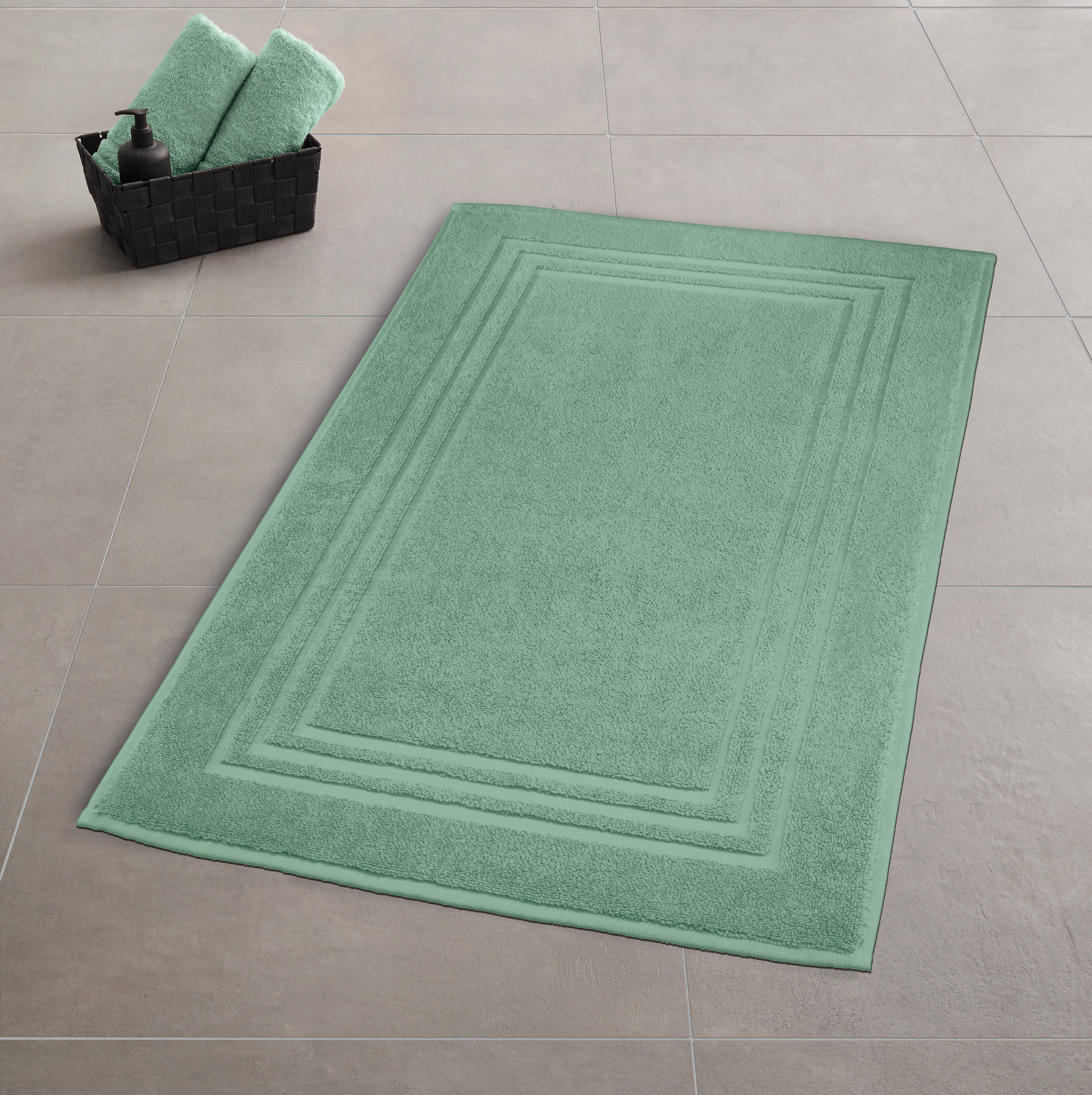 Grüner Badteppich auf grauem Fliesenboden, daneben ein schwarzer Korb mit grünen Handtüchern und einer schwarzen Seifenspenderflasche.