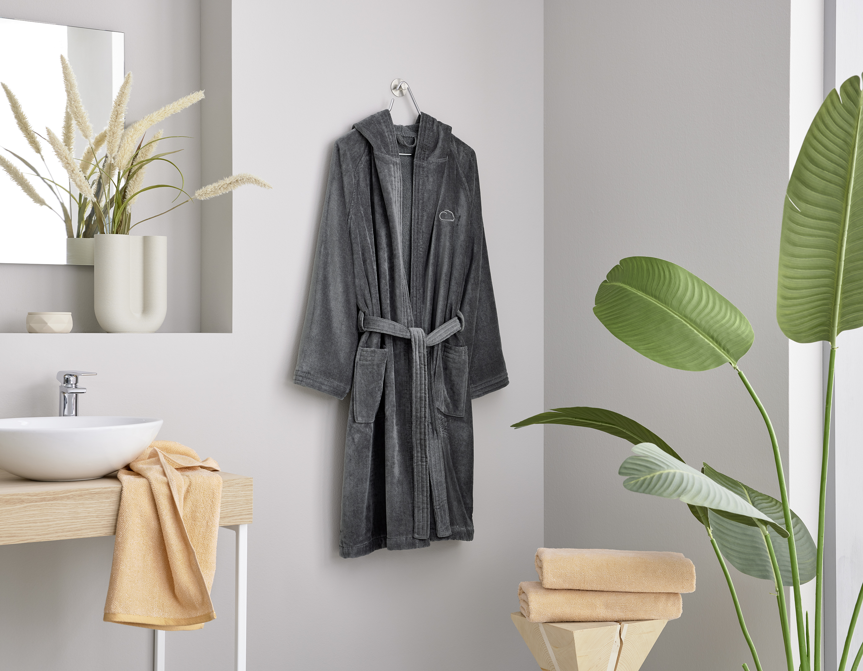 Grauer Bademantel an Haken, modernes Badezimmer mit Pflanzen, Waschbecken und Handtüchern in Beige und Gelb.