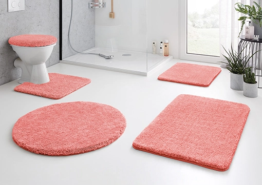 Korallfarbene Badematten-Set in modernem Badezimmer, bestehend aus WC-Deckelbezug, WC-Vorleger, Duschvorleger und zwei rechteckigen Matten.