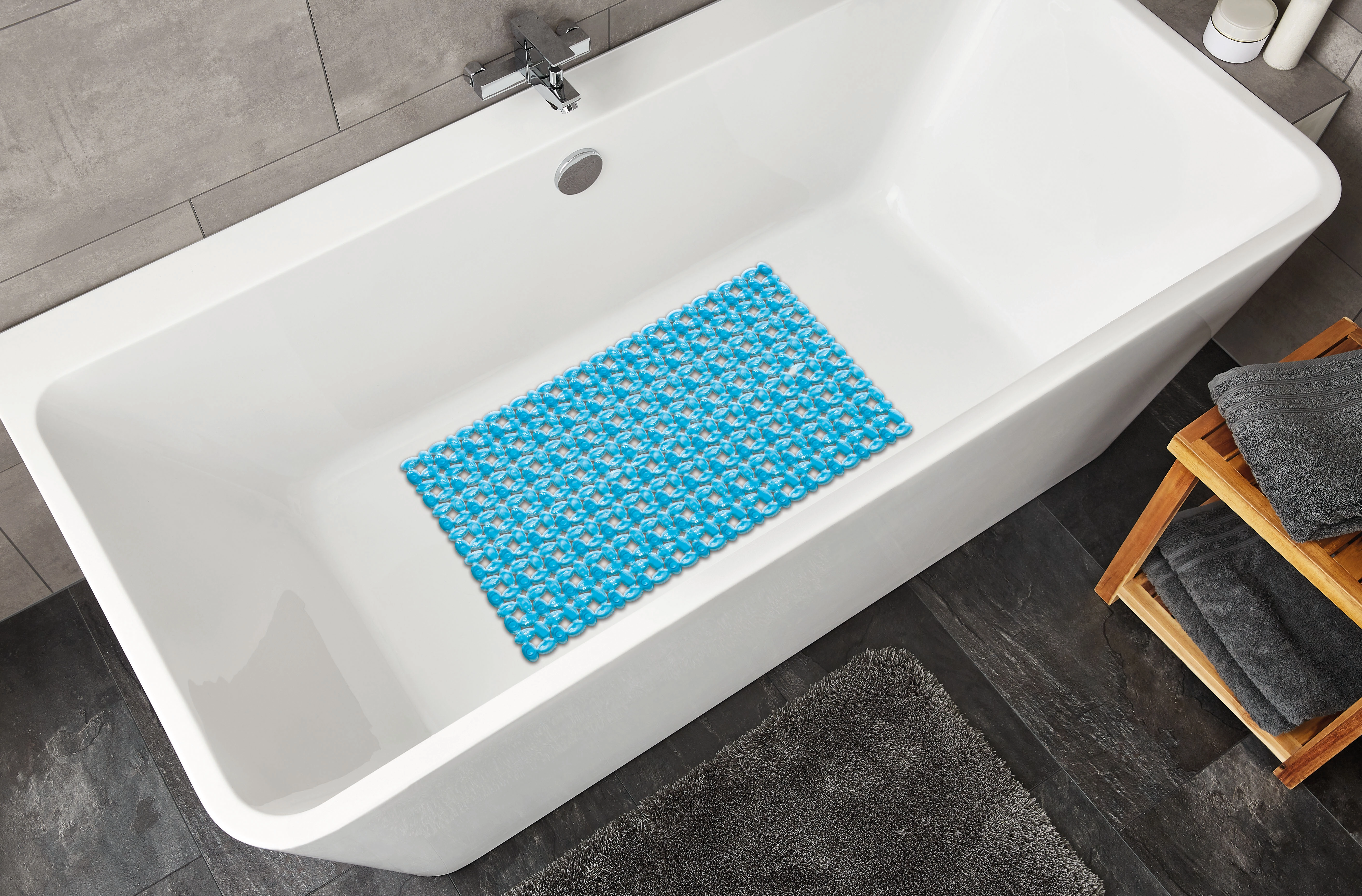 Blaue Badematte mit floralem Muster in einer modernen weißen Badewanne, umgeben von grauen Handtüchern und dunklen Fliesen.