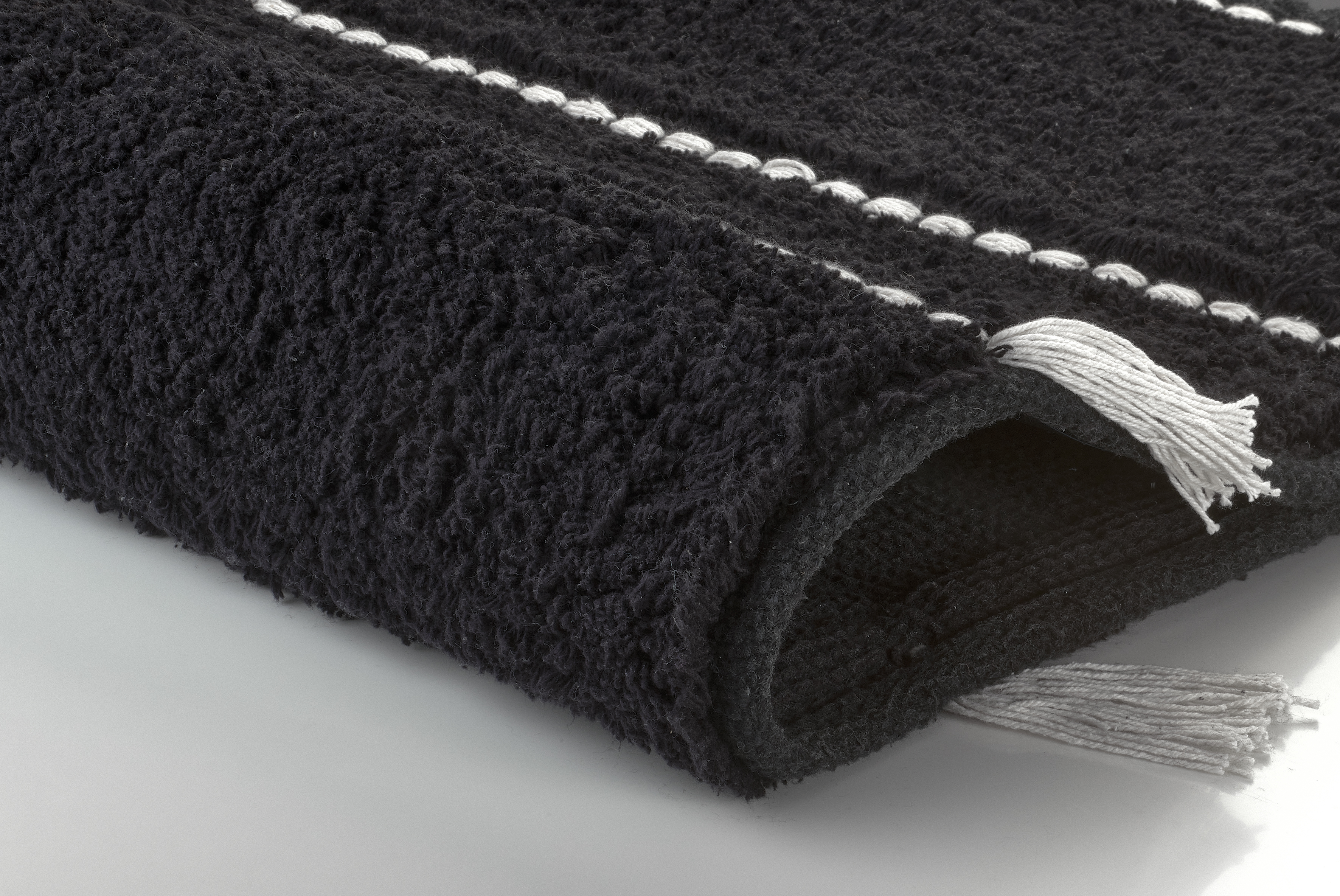 Schwarzer, flauschiger Badteppich mit weißen Streifen und Fransen, aufgerollt auf einer glatten Oberfläche.
