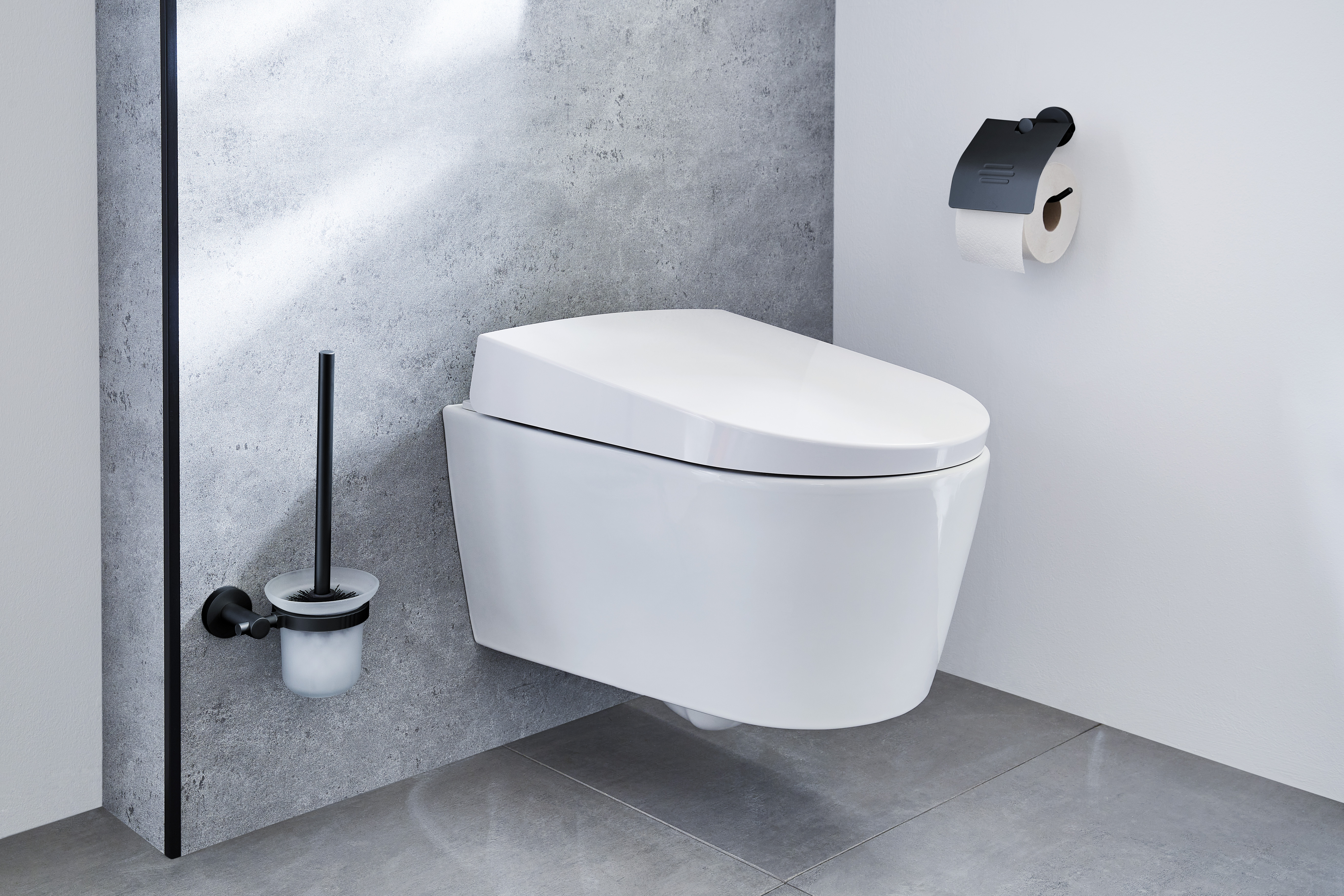 Modernes, wandhängendes WC in einem minimalistischen Badezimmer mit grauen Fliesen, Toilettenpapierhalter und WC-Bürste an der Wand.